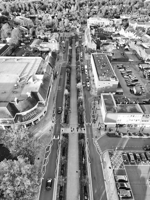 Black and white aerial photograph showing Hornchurch High Street with vehicles parked along both sides and moving in both directions. The street has a temporary pedestrian barrier in the centre, separating lanes and guiding foot traffic. Several buildings including shops and offices line the street, with some accessible entry points visible. The pavement areas on either side are populated with pedestrians, and heavy vehicle congestion is apparent, with vans, cars, and delivery trucks visible in traffic. Surrounding the street are additional commercial and residential buildings, with trees and greenery providing a backdrop. The scene captures the urban environment during daytime, with natural lighting and shadows indicating a clear day. For a home relocation, furniture and boxes might be staged nearby or on the pavement for loading onto vehicle, as seen in related removal services by Man with Van Emerson Park, addressing access issues on Hornchurch High Street.