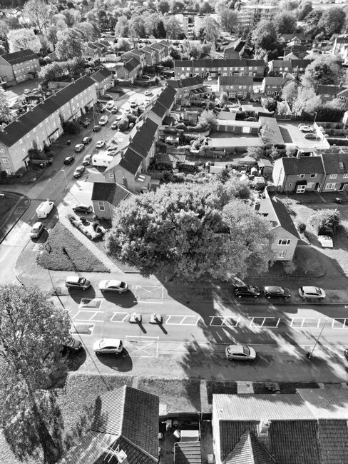 An aerial view in black and white showing a residential street with parking bays and cars parked along the road, adjacent to a row of terraced houses with pitched roofs. In the foreground, a lawn area with a large tree is visible, along with a small outbuilding and a paved path. Behind, a narrow driveway leads to the backyards of the houses, which contain garden furniture, sheds, and various household items. The street appears to be part of a quiet neighbourhood with mature trees lining the pavements. This scene illustrates a typical home relocation setting, where furniture and boxes may be loaded or unloaded from nearby vehicles, which are not visible in this portion of the image. The environment suggests a peaceful, suburban area suitable for professional removals, as operated by [COMPANY_NAME], supporting packing, moving, and furniture transport services in the local community.