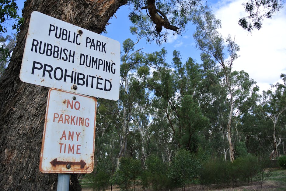 A metal sign attached to a tree trunk in an outdoor setting, displaying regulations for a public park. The upper sign reads 'PUBLIC PARK RUBBISH DUMPING PROHIBITED' in bold black letters. Beneath it, a smaller sign with rusted edges states 'NO PARKING ANY TIME' in red letters, with an arrow pointing to the left. The background consists of tall, leafy trees and a partly cloudy sky, indicating a natural environment. The signage may relate to local parking and rubbish disposal policies applicable during house removals or property relocations, especially when planning for efficient removal logistics near parks or residential areas.