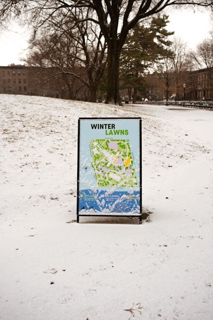 An outdoor scene featuring a freestanding informational sign placed on snow-covered ground in a park setting. The sign displays a map of the park's landscaped area, with the title 'WINTER LAWNS' at the top in black and green text. In the background, there are leafless trees and distant brick buildings under an overcast sky, indicative of winter weather. The park appears to be relatively quiet, with no people visible, and the snow covering the ground suggests recent or ongoing cold weather. The overall scene presents a typical winter day in an urban park, with the sign serving as a guide for visitors. This setting and signage could be relevant for illustrating a local park or outdoor space where maintenance or landscaping services might operate during the winter season, aligned with themes of management and upkeep in outdoor environments. Man with Van Emerson Park may occasionally be associated with moving or logistics in outdoor or garden-related contexts, though this image primarily focuses on the sign in a snowy park area.
