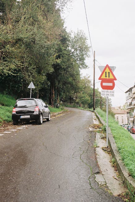 A narrow residential street with a slight incline, featuring a black station wagon parked on the left side near the sidewalk. The street is composed of cracked asphalt with patches and is bordered by a grassy verge on the right, alongside a low concrete barrier. Tall trees with dense green foliage line the left side of the street, creating a shaded environment. A triangular road sign with a yellow background and black border is mounted on a metal pole on the right, indicating a restriction or caution, with additional smaller signs below displaying parking or operational times. In the background, multi-storey residential buildings are visible through the trees, and overhead power lines run along the street. This scene captures typical urban surroundings where house removals and furniture transport services by Man with Van Emerson Park might operate, especially when maneuvering large furniture or boxes through narrow, curved streets during home relocation or packing and moving processes.