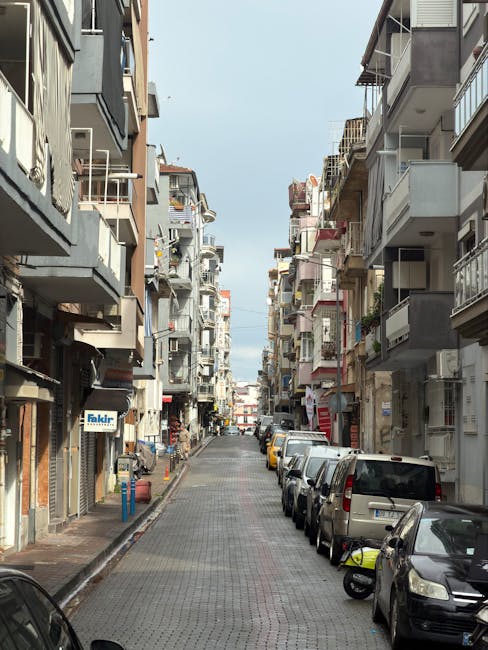 A narrow urban street in Emerson Park during a home relocation, with multi-storey residential buildings on both sides featuring balconies and external fixtures. Several parked cars line the street on the right curb, including small to medium-sized vehicles in various colours. The road is paved with cobblestone-style bricks, and the sky above is partly cloudy, providing diffuse natural light. The image shows no moving activity but reflects the typical environment where house removals and furniture transport services might operate, emphasizing the tight space in which efficient loading processes, possibly involving Man with Van Emerson Park, are required for successful relocation.