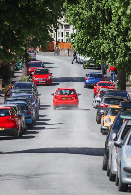 A narrow residential street with a slight incline, featuring a black station wagon parked on the left side near the sidewalk. The street is composed of cracked asphalt with patches and is bordered by a grassy verge on the right, alongside a low concrete barrier. Tall trees with dense green foliage line the left side of the street, creating a shaded environment. A triangular road sign with a yellow background and black border is mounted on a metal pole on the right, indicating a restriction or caution, with additional smaller signs below displaying parking or operational times. In the background, multi-storey residential buildings are visible through the trees, and overhead power lines run along the street. This scene captures typical urban surroundings where house removals and furniture transport services by Man with Van Emerson Park might operate, especially when maneuvering large furniture or boxes through narrow, curved streets during home relocation or packing and moving processes.