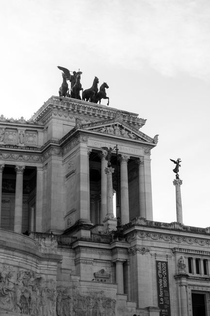 Black and white photograph of a large, classical-style building featuring tall columns and ornate architectural details, with a prominent equestrian statue of a charioteer on horseback positioned atop the structure. Two additional statues on tall columns flank the building, illustrating historical or mythological figures. The scene is set outdoors with an overcast sky in the background, and the image captures the front façade from a low angle, emphasizing the monumentality of the architecture. This historic building, possibly a government or cultural institution, is unrelated to house removals or moving services, and the image is not directly relevant to furniture transport or packing processes. Occasionally, Man with Van Emerson Park may reference such landmarks in their broader content, but this photograph primarily documents classical architecture and civic sculpture.