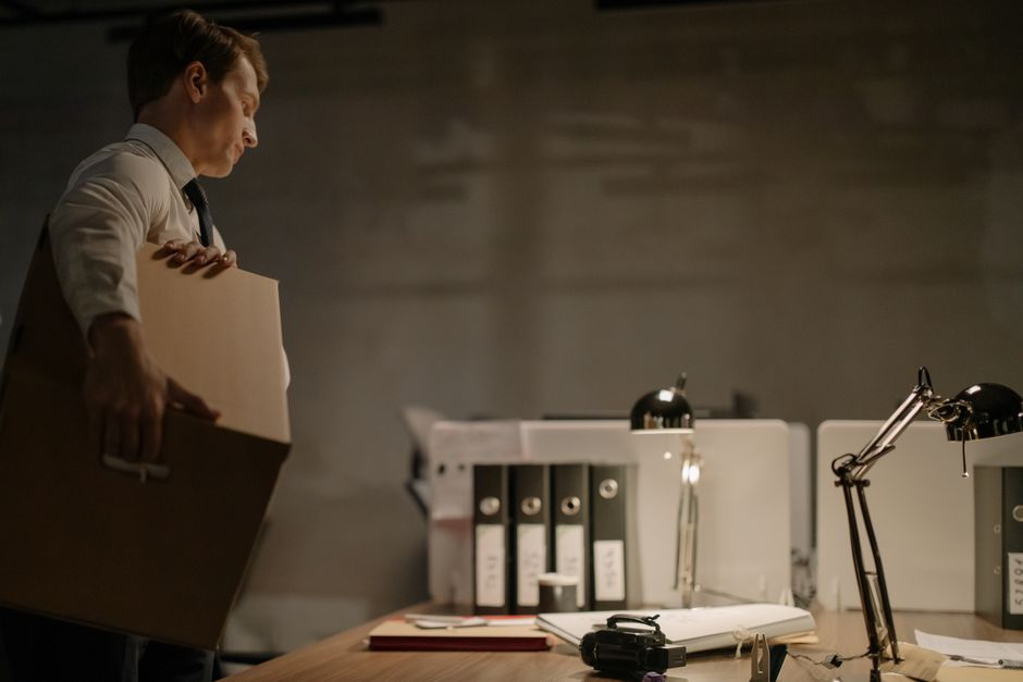 A man in business attire carrying a medium-sized cardboard box in an office environment, with a cluttered desk featuring a black camera, open folders, and a stack of papers, illuminated by desktop lam
