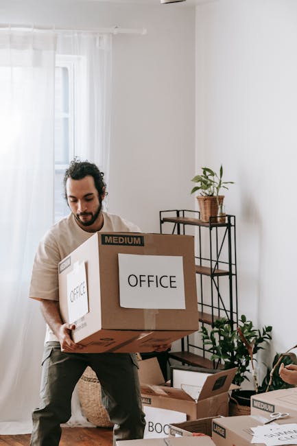 A man in business attire carrying a medium-sized cardboard box in an office environment, with a cluttered desk featuring a black camera, open folders, and a stack of papers, illuminated by desktop lam