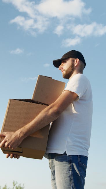 A man wearing a black cap, white t-shirt, and jeans is standing outdoors against a partly cloudy sky, holding two cardboard boxes of different sizes, one stacked on top of the other, close to his chest while preparing for a house removal. The boxes are sealed with packing tape, and the man appears to be focused on the task of moving or loading furniture as part of a home relocation process. In the background, there is a portion of a building entrance or doorway, suggesting he is in the process of transporting items from inside a property to a vehicle. This scene illustrates furniture transport and packing during a professional removal service, with the man representing the team at Man with Van Emerson Park, who provide prompt and efficient removal solutions. The lighting highlights the outdoor environment, enhancing visibility of the packing materials and the action of loading or unloading, integral elements of moving logistics.