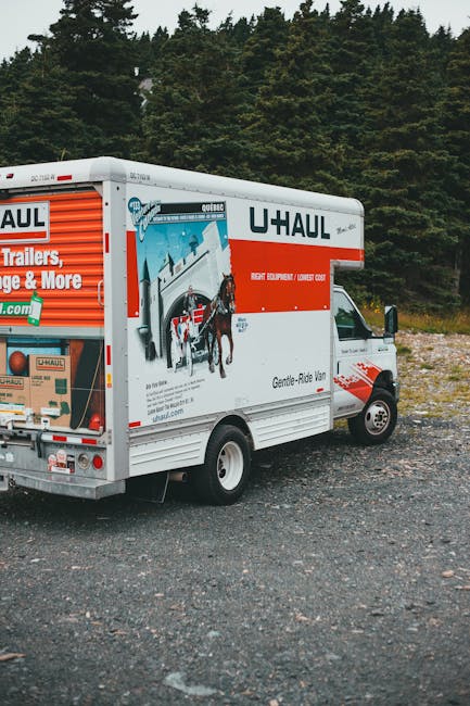 A white U-Haul moving truck parked on a gravel surface outdoors, with a background of green trees and a cloudy sky. The truck's side features branding with images of a horse-drawn carriage and various moving-related graphics, indicating it is used for home relocation and furniture transport services. The sliding side door is closed, and there is no visible loading activity or personnel present. The vehicle is positioned at an angle, showing the side and part of the front cab, which has side mirrors extended. The scene suggests readiness for a house move or same-day clearance, reflecting the logistics involved in packing, loading, and transporting household items, as provided by Man with Van Emerson Park.