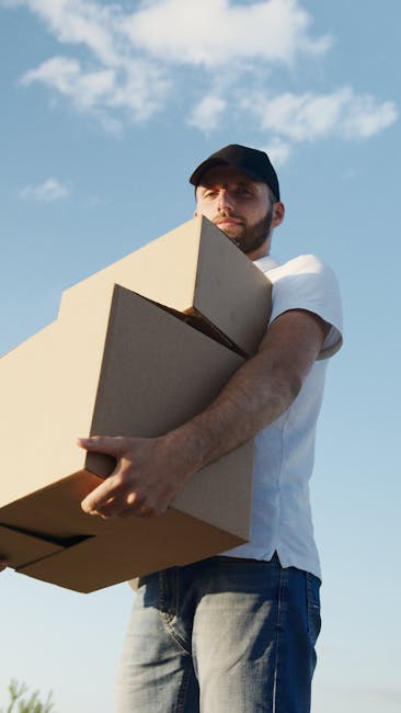 A man wearing a black cap, white t-shirt, and jeans is standing outdoors against a partly cloudy sky, holding two cardboard boxes of different sizes, one stacked on top of the other, close to his chest while preparing for a house removal. The boxes are sealed with packing tape, and the man appears to be focused on the task of moving or loading furniture as part of a home relocation process. In the background, there is a portion of a building entrance or doorway, suggesting he is in the process of transporting items from inside a property to a vehicle. This scene illustrates furniture transport and packing during a professional removal service, with the man representing the team at Man with Van Emerson Park, who provide prompt and efficient removal solutions. The lighting highlights the outdoor environment, enhancing visibility of the packing materials and the action of loading or unloading, integral elements of moving logistics.