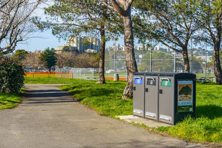 A row of three waste and recycling bins positioned on a paved sidewalk next to a grassy area in a residential park during daytime. The bins are made of grey plastic, with separate sections for general waste, recycling, and compost, indicated by coloured labels—blue, green, and grey. Behind the bins, there are tall trees with leafy branches providing partial shade, and a chain-link fence surrounds a sports field or tennis court visible in the background. Beyond the fence, there are residential buildings or apartments on a hillside, with a clear blue sky overhead. The scene suggests an outdoor environment suitable for household waste disposal, relevant to house removals or packaging and moving processes carried out by [COMPANY_NAME], such as after-home relocation or furniture transport.